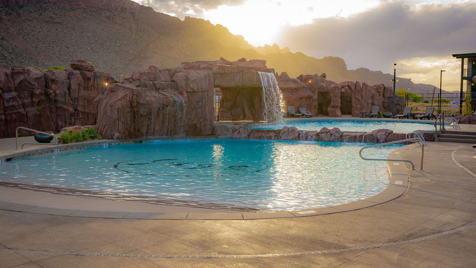 Large luxury swimming pool with rock waterfall and sun setting in the background - Precision Pools